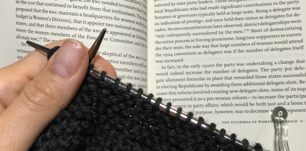 Black yarn being knitted with an open book in the background.